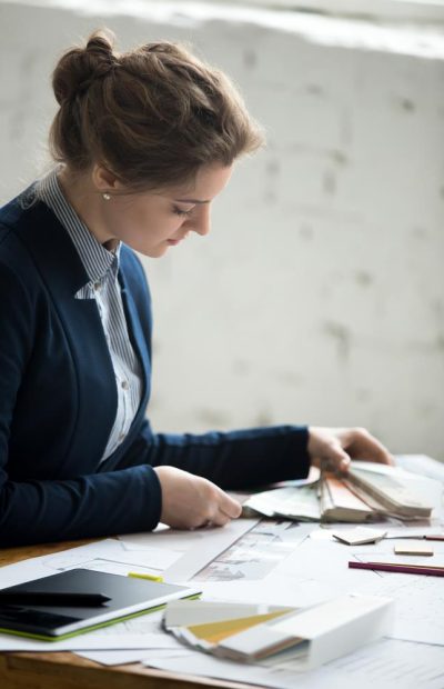 Portrait of beautiful young designer woman wearing suit working at new project with drafts and color swatches at office desk. Attractive model choosing material samples. Interior shot
